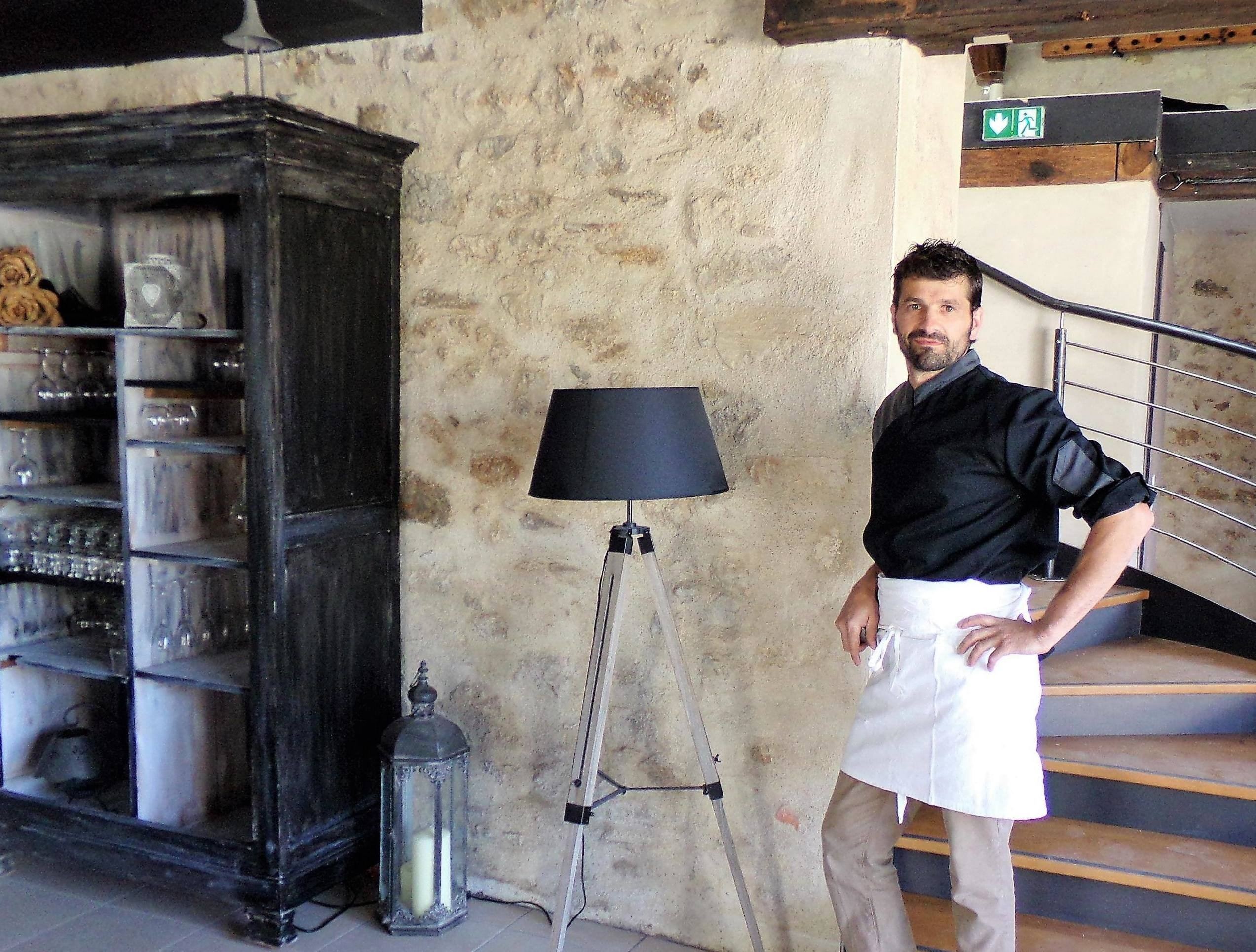 Le chef en veste noire et tablier blanc, dans la salle aux murs de pierre