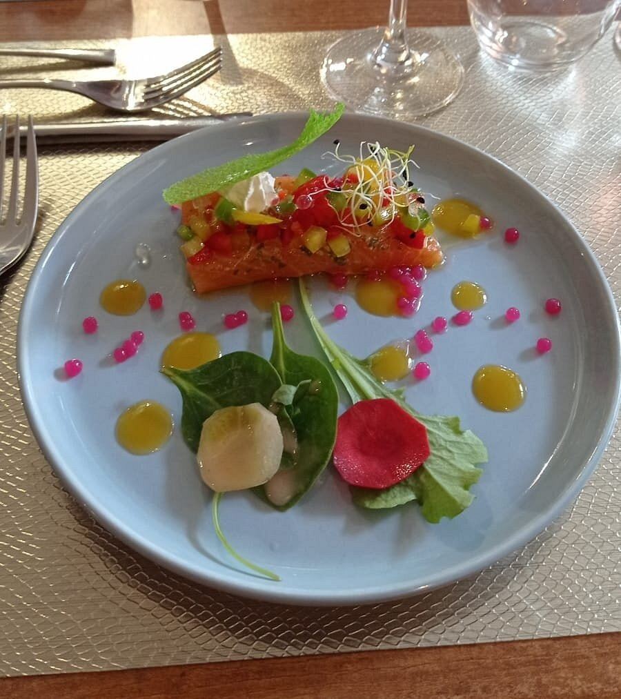 Pavé de saumon surmonté de petits légumes en dés, perles roses, points de sauce jaune et jeunes pousses, dans une assiette gris-bleu